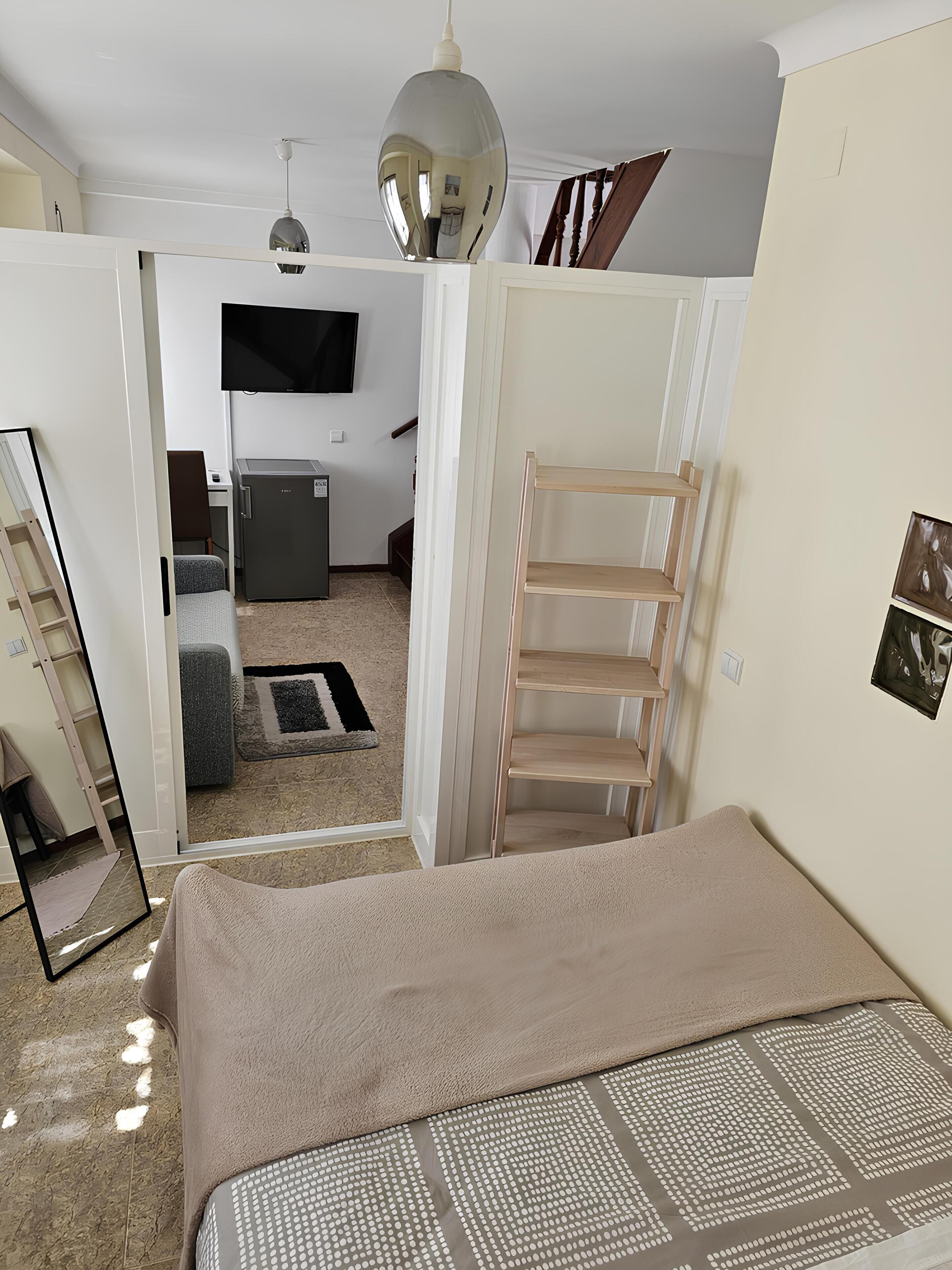 Un dormitorio con cama, armario, escritorio, silla, espejo, televisor y un mueble pequeño.