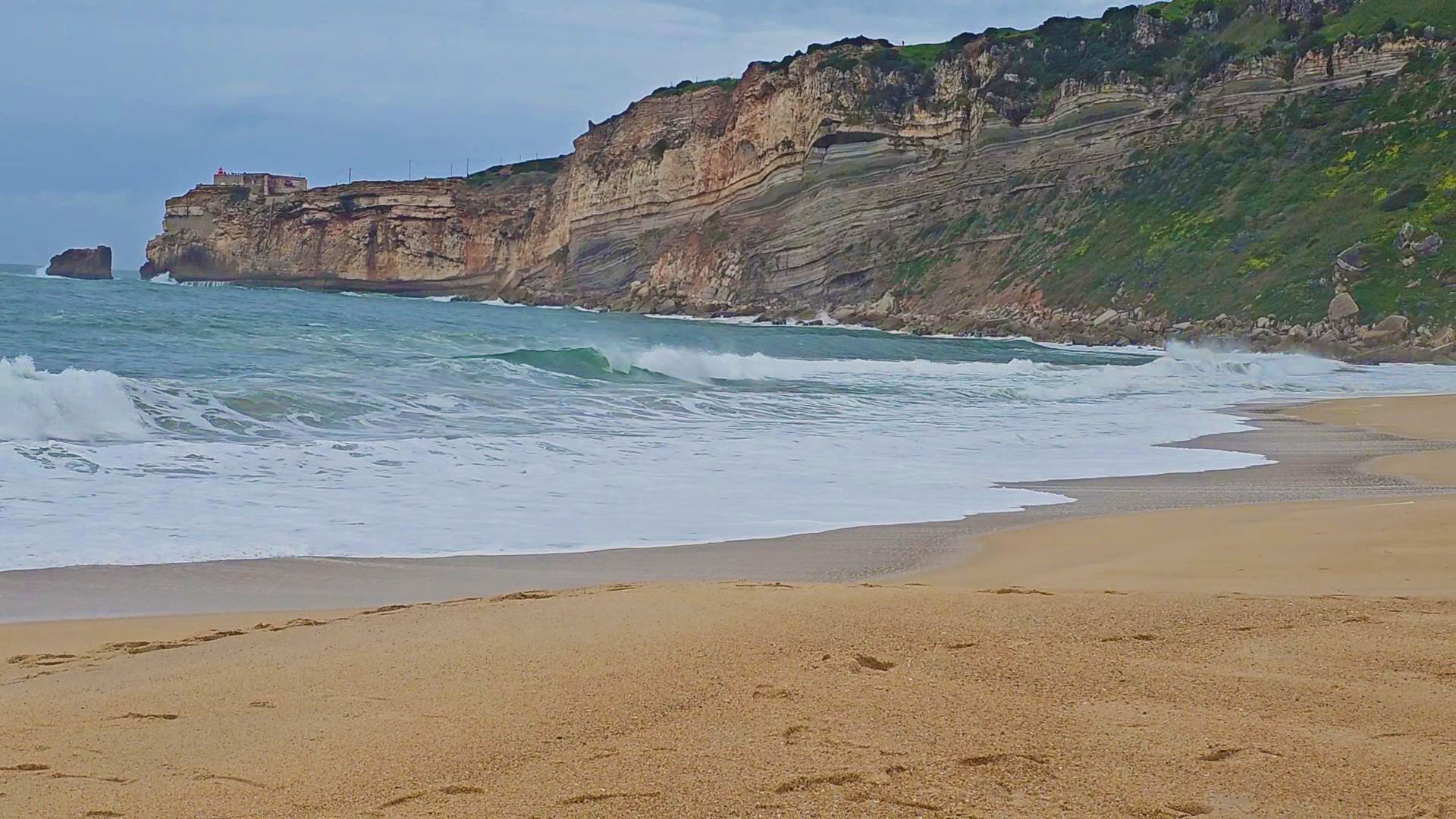 Ein Strand mit brechenden Wellen, eine Klippe mit schichtartigen Felsformationen und eine Struktur auf der Klippenkuppe.