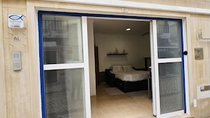 A hotel entrance with a glass door, a bed with a patterned bedspread, and a small shelf with decorative items. -  Sunao (Nazaré)