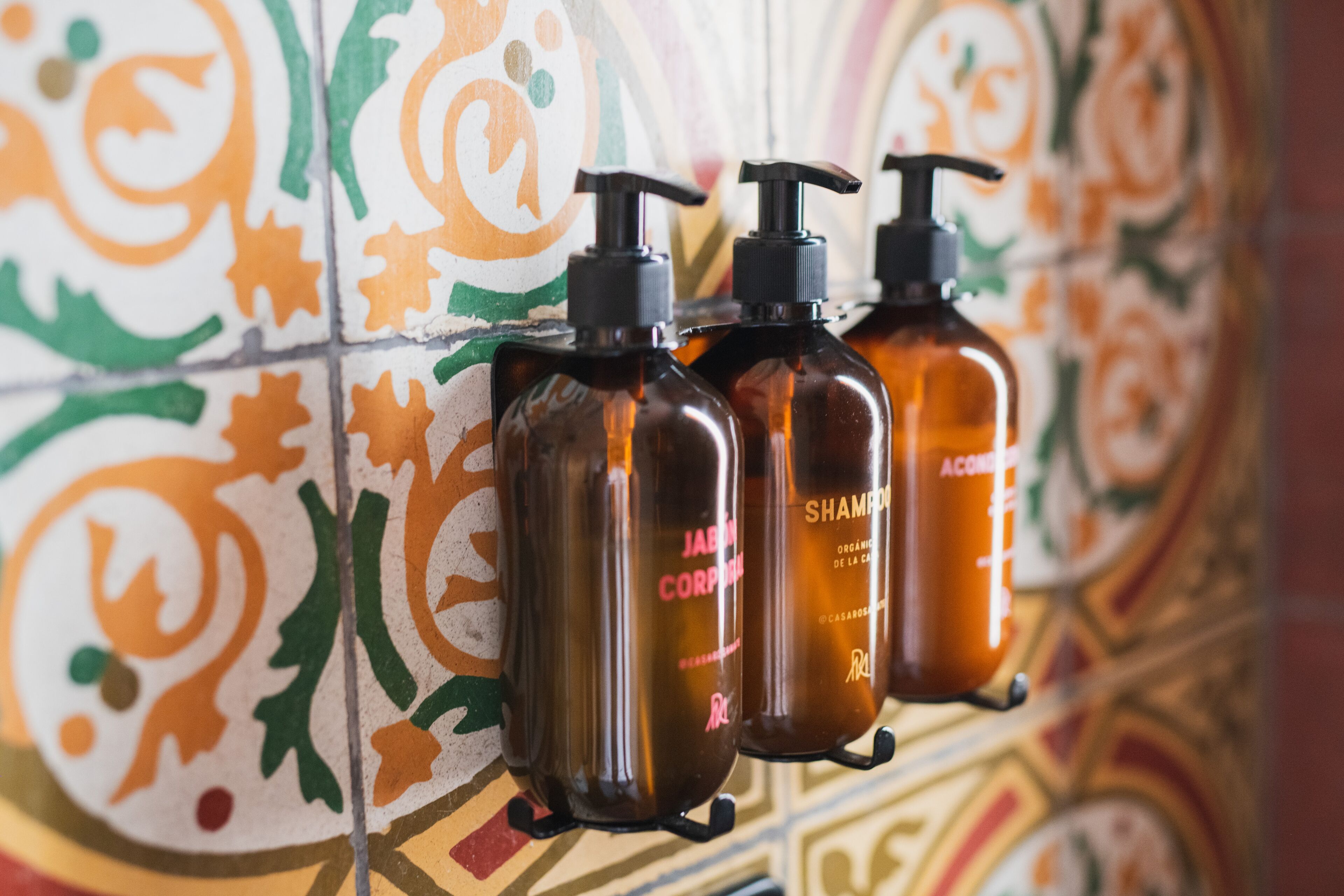 Tres amples flaços marrons amb polsadores negres etiquetats com 'JABON CORPORAL' i 'XAMPÚ' davant d'una paret de rajoles de colors.