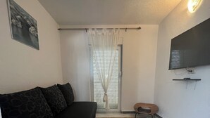 Uma sala de estar com um sofá preto, um tapete tecido, uma televisão na parede e uma janela com cortinas leves.