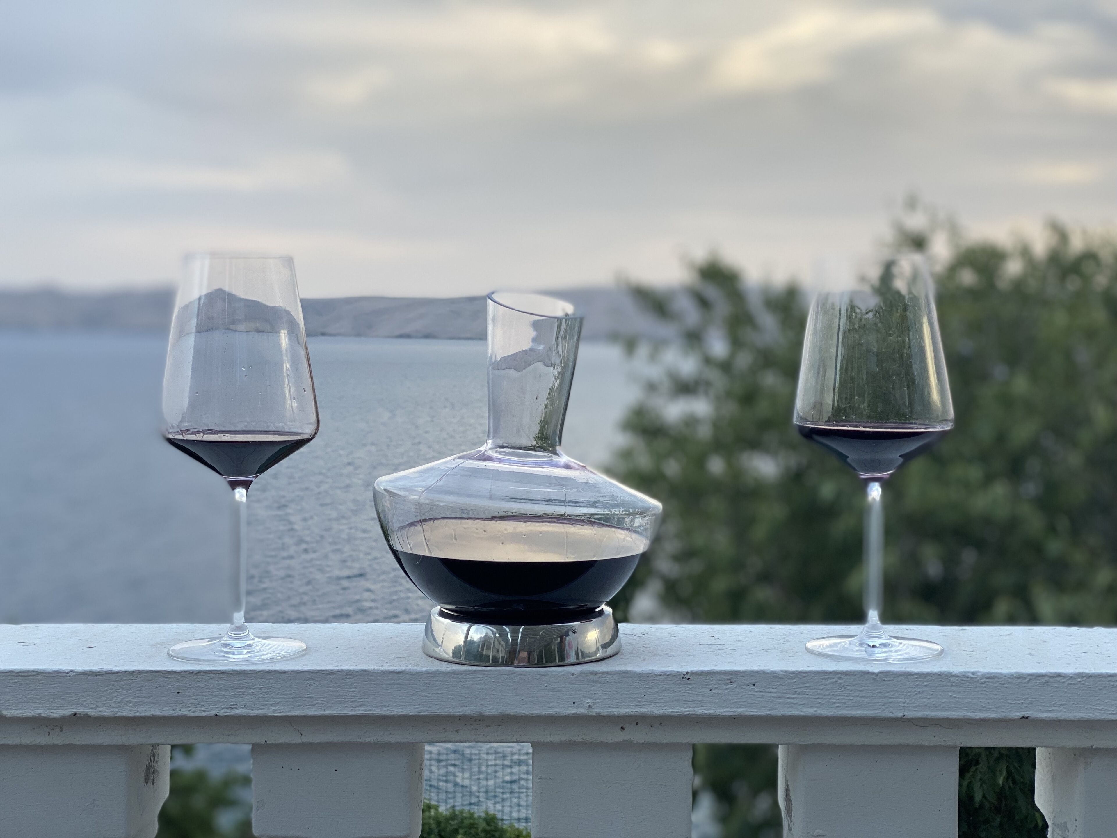 Three wine glasses and a decanter on a white railing with a view of the sea and hills in the background.