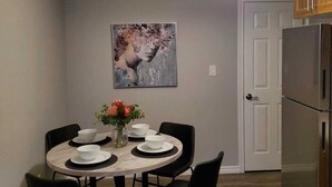 Un comedor con una mesa redonda, sillas negras y un jarrón con flores. Hay un cuadro en la pared y un refrigerador al fondo.