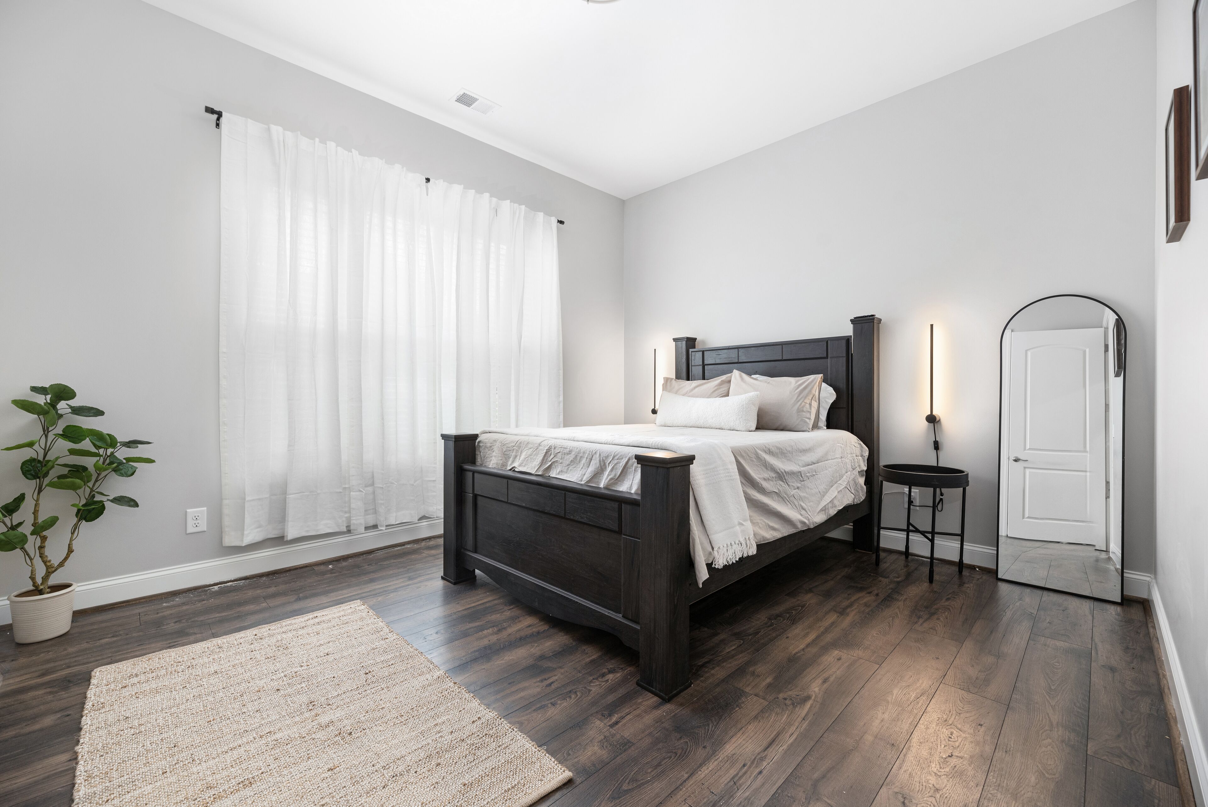 A bedroom with a dark wooden bed, white bedding, a bedside table with a lamp, a potted plant, and a large window with white curtains.
