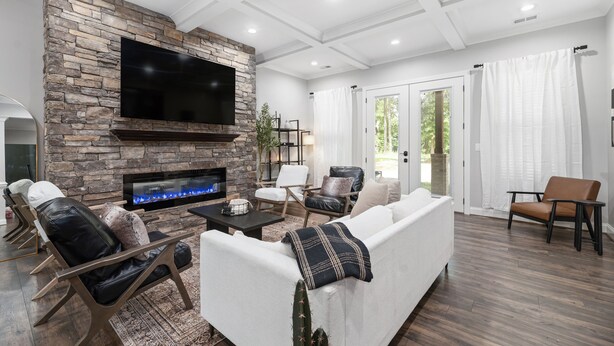 Una sala de estar moderna con una chimenea de piedra, un televisor de pantalla plana, sillones de cuero, un sofá blanco y un ventanal con cortinas.