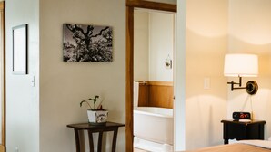 Ein Hotelzimmer mit Bett, Nachttisch mit Lampe, kleinem Tisch mit Topfpflanze, gerahmtem Bild an der Wand und Spiegel, der das Badezimmer reflektiert.