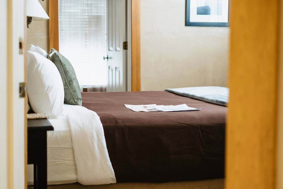 A neatly made bed with a brown bedspread and white pillows, a bedside table with a lamp, and a door leading to a room with a window.