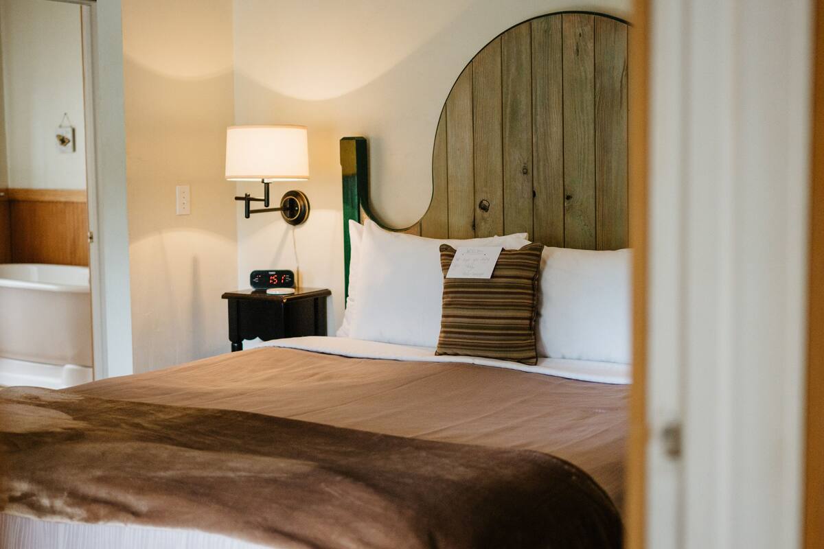 A hotel room with a large bed, a wooden headboard, a nightstand with a lamp, and a bathroom visible through an open door.