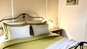 Ein ordentlich bezogenes Bett mit einer grün-weißen Tagesdecke, einem schwarzen Metall-Kopfteil und einem Nachttisch mit einer Lampe und einer kleinen Pflanze.