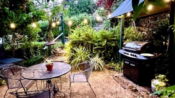 Eine Terrasse mit rundem Tisch, Stühlen, üppiger Bepflanzung und einem Grill.