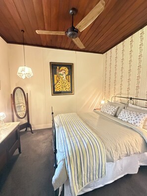 A bedroom with a bed, a ceiling fan, a framed picture, a mirror, and patterned wallpaper.