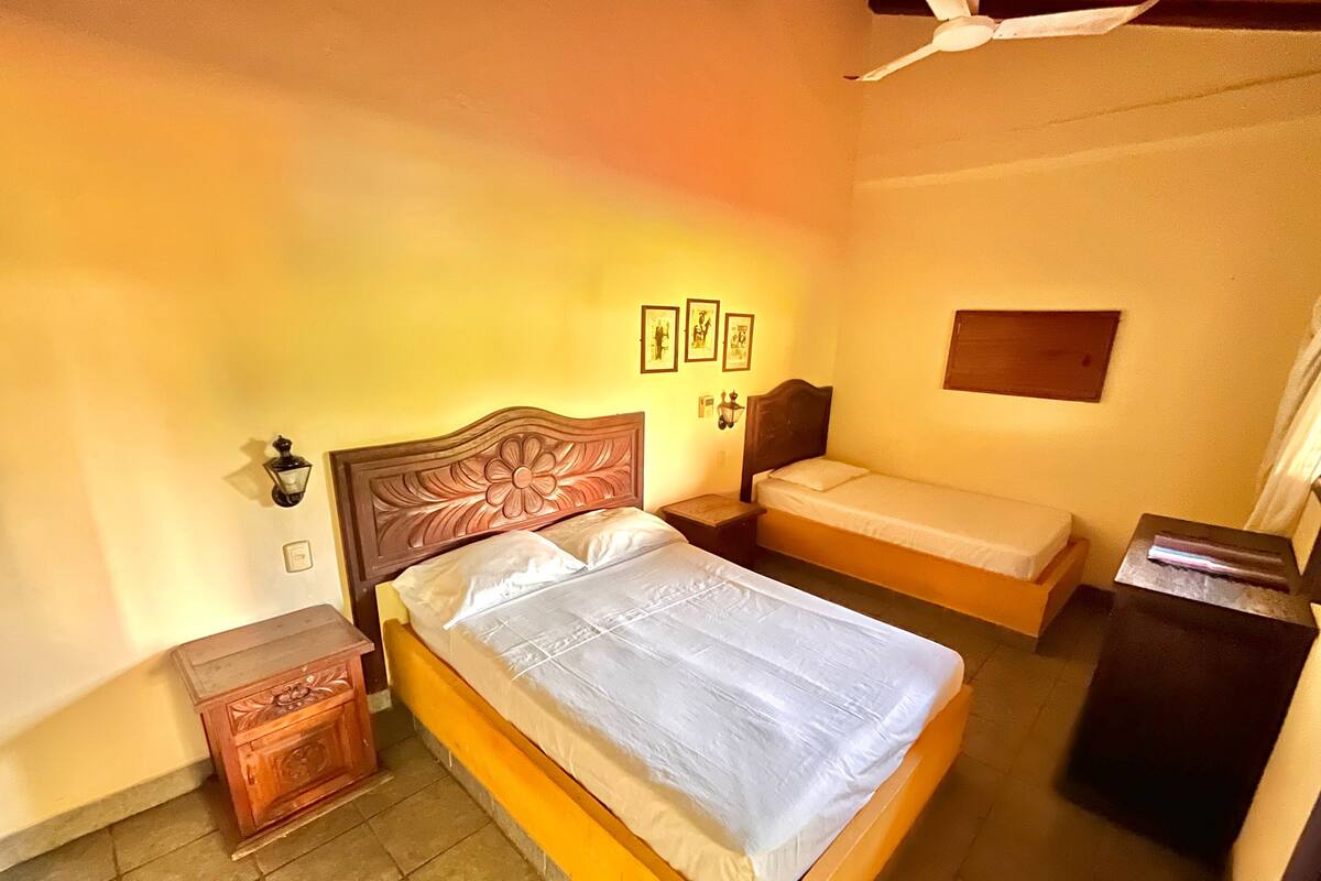 A bedroom with a large bed, a smaller bed, a nightstand, a cabinet, a wall-mounted lamp, and framed pictures on the wall.