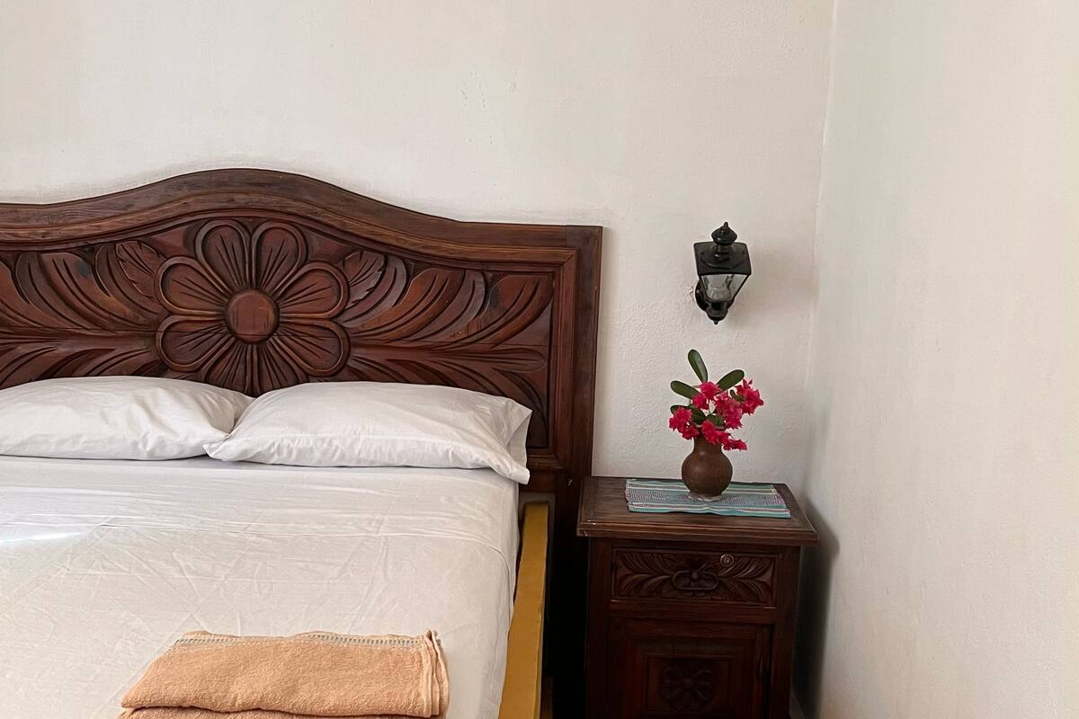 A neatly made bed with white linens and a folded beige towel, a wooden nightstand with a vase of pink flowers, and a wall-mounted light fixture.