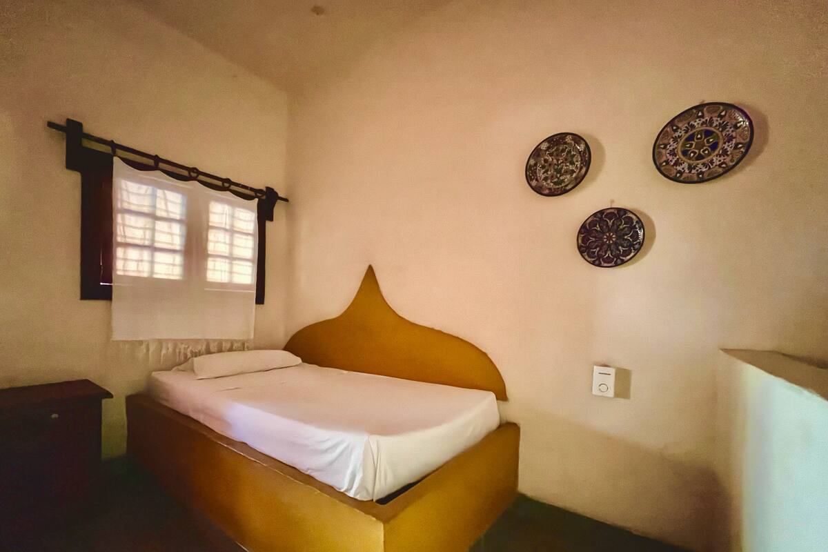 A single bed with a wooden headboard in a room with plain walls. There are two wall-mounted decorative plates and a window with a curtain.