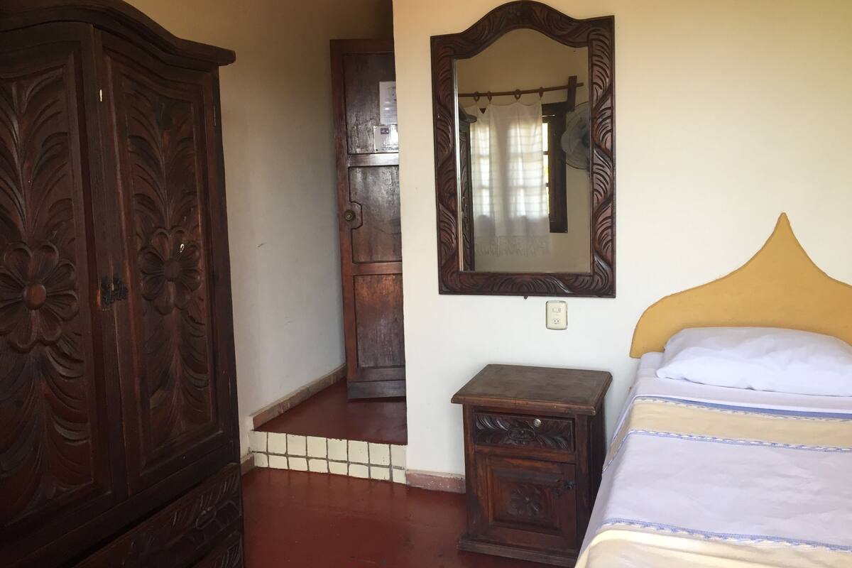 A bedroom with a wooden wardrobe, a bedside table, a mirror, and a bed with a yellow headboard.