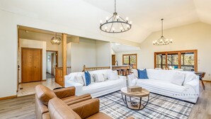 A spacious living room with a large white sectional sofa, a brown leather couch, and a round coffee table. There are two chandeliers hanging from the ceiling, and a wooden staircase leading to another level. - Partridge Place | 4 Ensuite Bedrooms, Hot Tub, Fire Pit, Dog-Friendly (Galena)