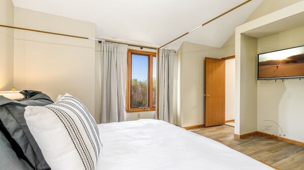 Une chambre avec un grand lit, une fenêtre avec des rideaux, une porte en bois et un téléviseur fixé au mur.