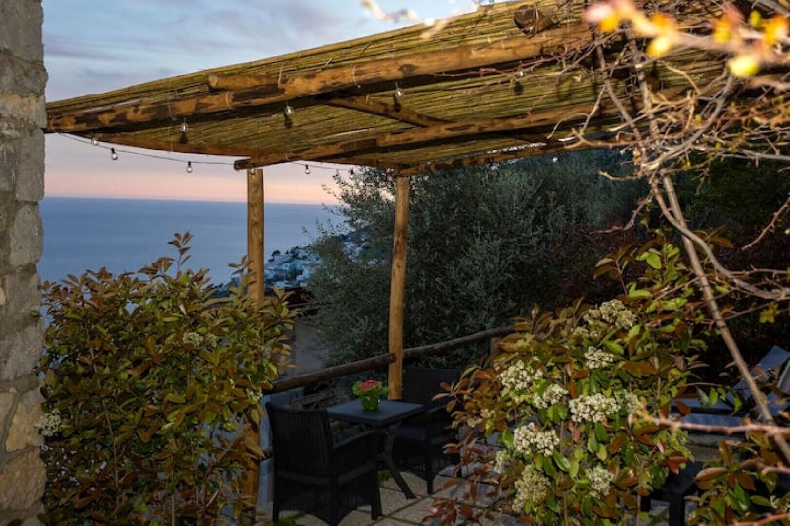A wooden pergola with string lights, a table and chairs, and a view of the sea.