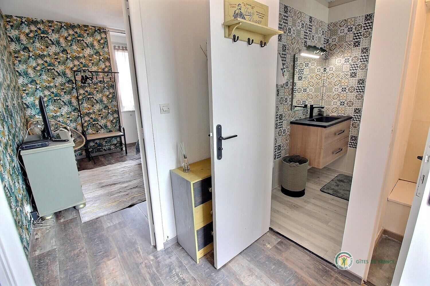 A room with a wooden floor, a small cabinet, a door with a towel rack, and a bathroom with patterned tiles and a sink.