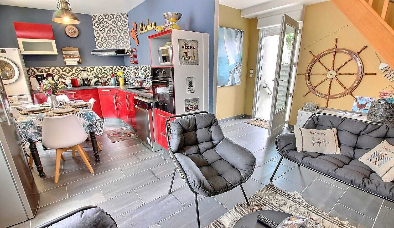 A modern kitchen with red cabinets, a dining area with a table and chairs, and a living room with a sofa and a ship's wheel decoration.