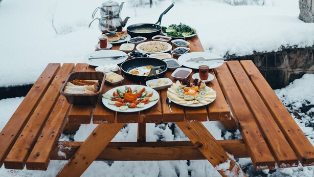 A wooden picnic table set up in a snowy landscape with various dishes and a teapot. - Tuğra Butik Otel (Savsat)