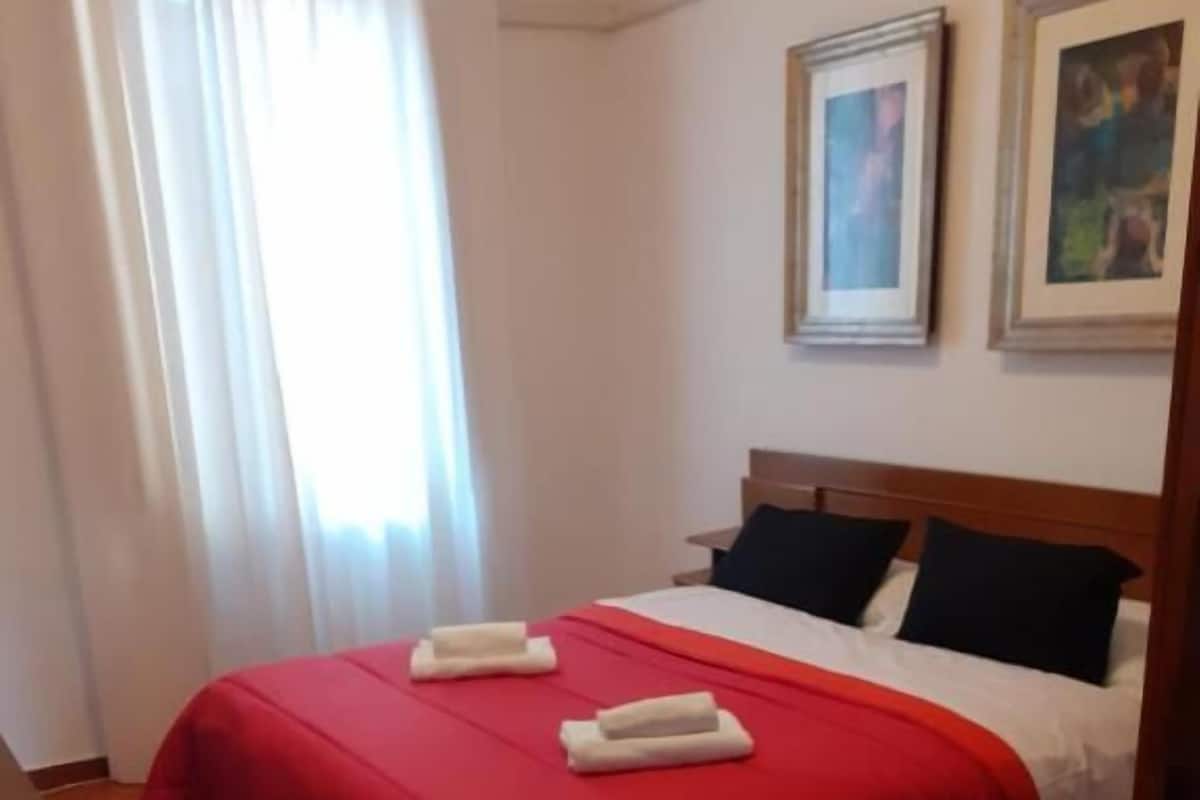 A hotel room with a bed, two pillows, a red bedspread, and a window with curtains.