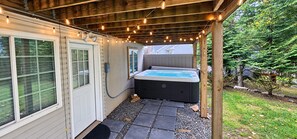 A hot tub under a wooden pergola with string lights, a door, and a stone pathway leading to a grassy area.