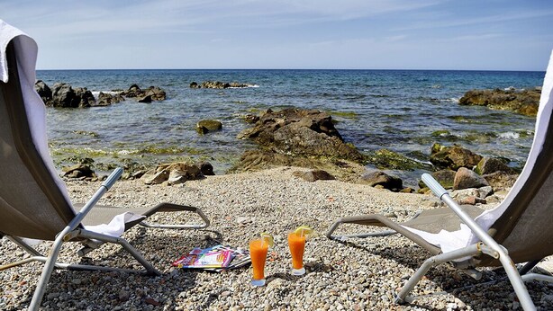 Två strandstolar placerade med ryggen mot havet, med en stenstrand i bakgrunden och en tidning på sanden.