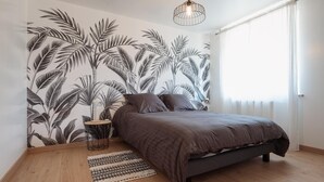 A bedroom with a bed, a nightstand, a lamp, a patterned rug, and a wall with a tropical leaf mural. - La Maison des Délices (DOSCHES)