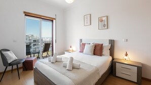 Une chambre d’hôtel moderne avec un grand lit, une chaise, une table de chevet avec une lampe et une vue sur la ville depuis les portes du balcon.
