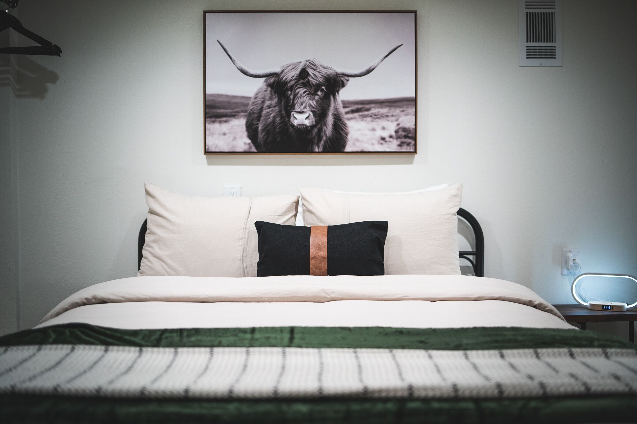 A bedroom with a bed, pillows, a blanket, a bedside table with a lamp, and a wall-mounted picture of a bull.