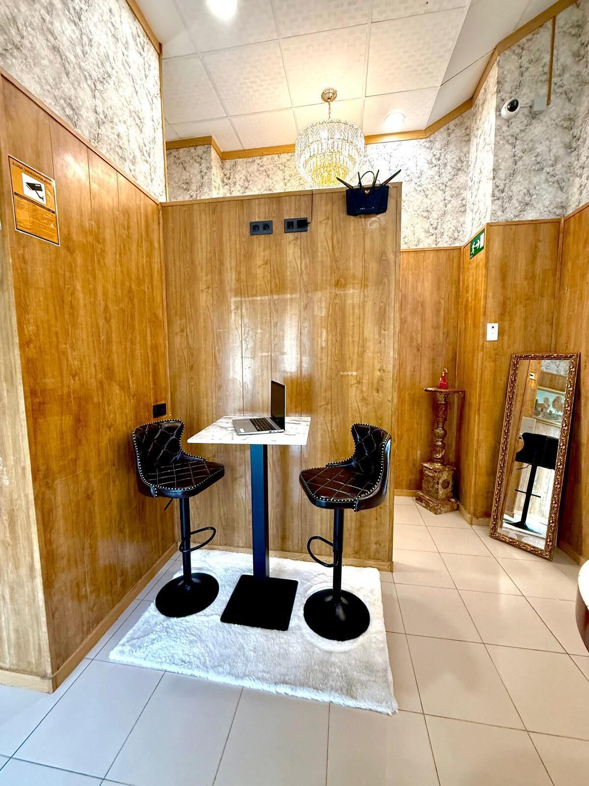 Una habitación con pared de madera, un candelabro, una mesa con una computadora portátil, dos sillas, un espejo y una pequeña estatua.