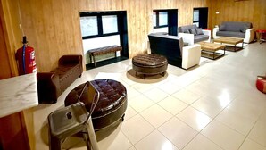 Amplia sala de estar con techo alto, paredes revestidas de madera y suelo de baldosas. Varias opciones de asiento, incluyendo un sofá, sillones y otomanas. Un extintor está montado en la pared.
