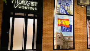 Exterior de Velvet Hostels con un letrero iluminado grande, un ventanal que muestra la bandera española y un muro de ladrillo.