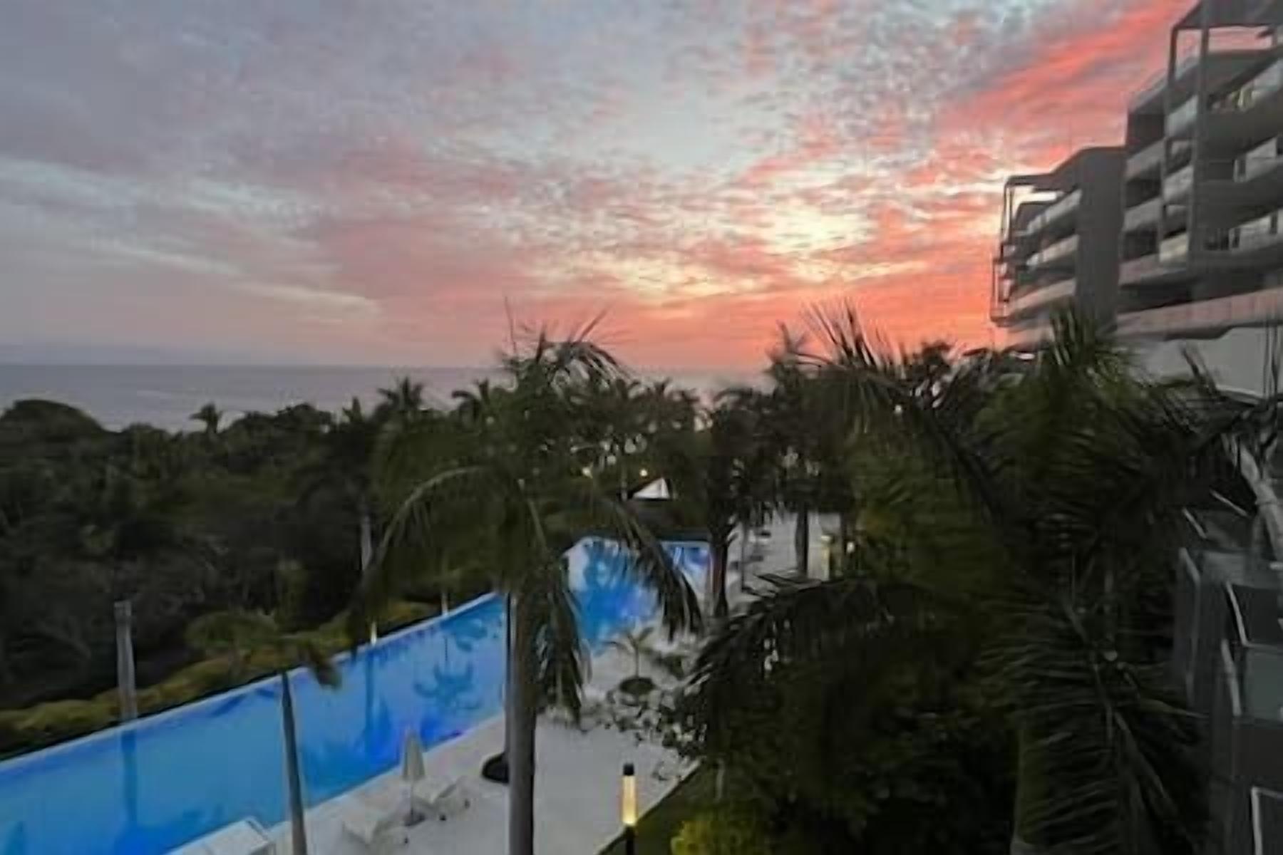A view of a coastal area with a sunset, palm trees, and a building with balconies.