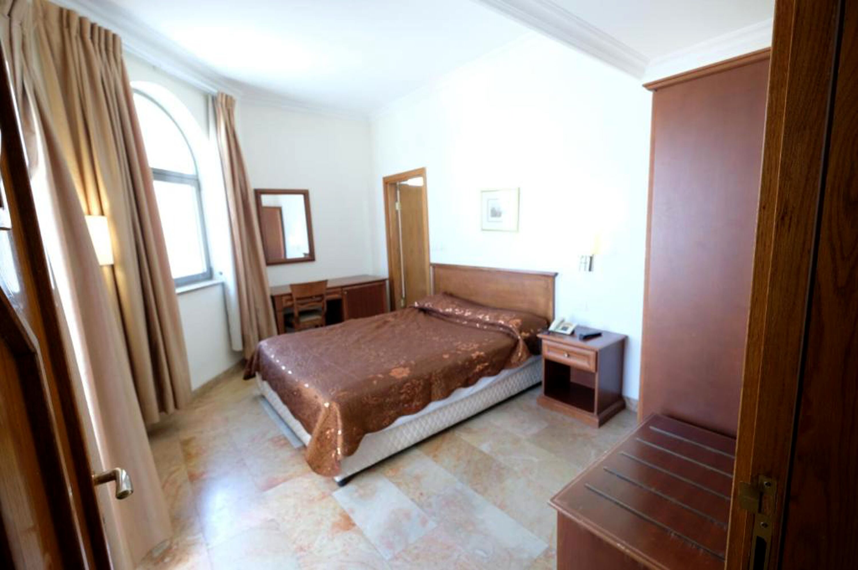 A bedroom with a bed, a nightstand, a wooden wardrobe, a mirror, and a window with curtains.