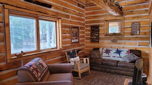 Interior de una cabaña de madera con sofá, sillón, mesa de madera, televisor y ventana con persianas.