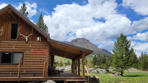 Una cabaña rústica de troncos con un porche, un paisaje montañoso de fondo y una decoración con cráneo de venado.