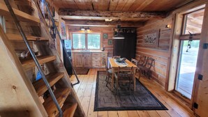 Interior rústico de una cabaña con una escalera de madera, un comedor con mesa y sillas, una cocina con armarios de madera y un gran ventanal que proporciona luz natural.