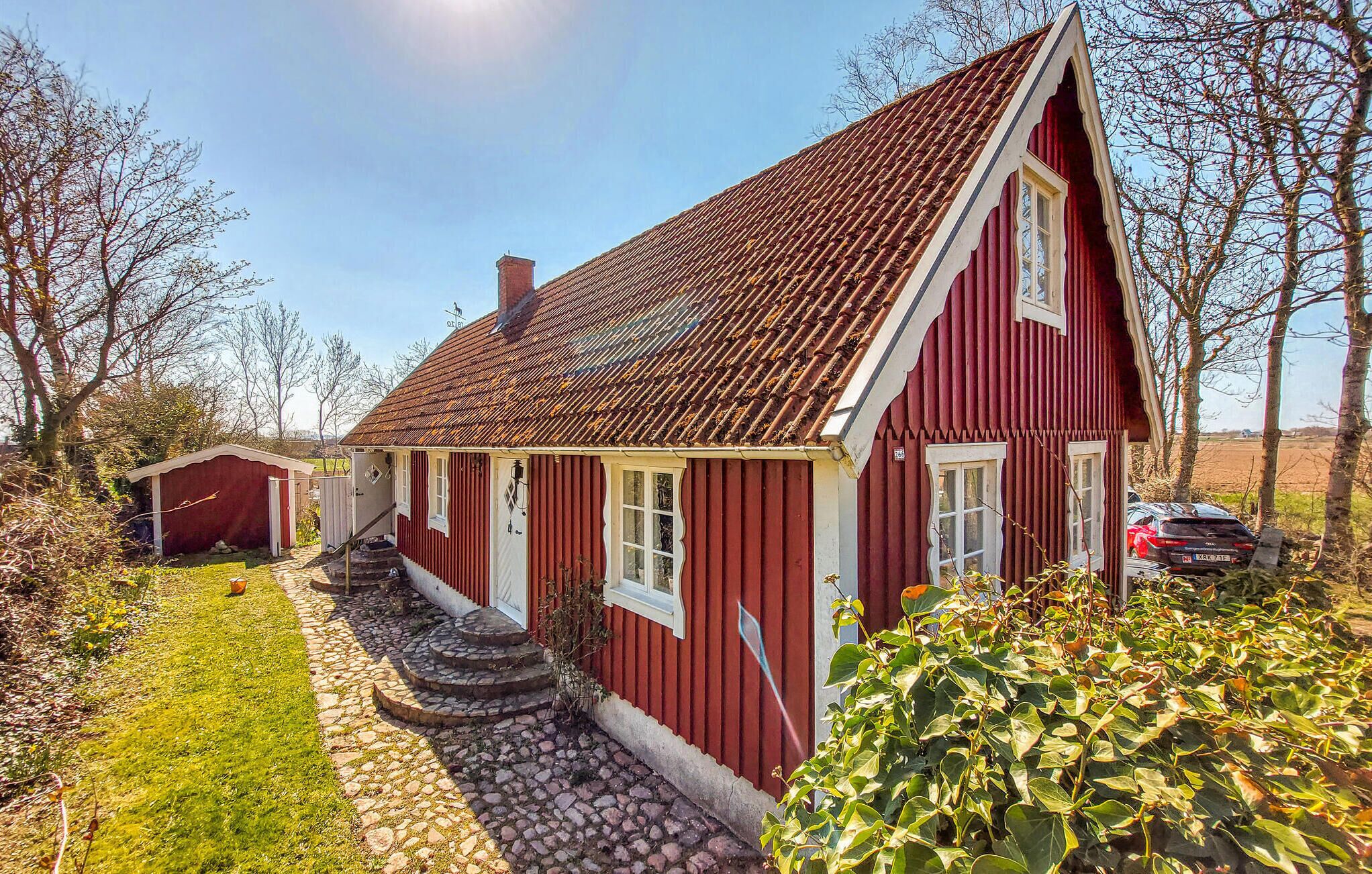 A red wooden house with a tiled roof, a stone staircase leading to the entrance, and a car parked in the driveway.