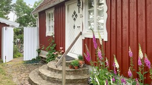 Une maison en bois rouge avec une porte et des fenêtres blanches, un escalier en pierre menant à la porte, ainsi que diverses plantes et fleurs en pot disposées autour de l’entrée.