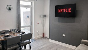 A modern living room with a dining area, a large flat-screen TV mounted on the wall, and a view through a large window. - Emerald Living - Swansea (Swansea)