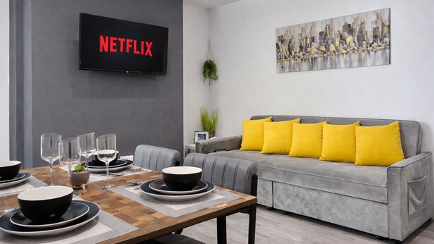 A modern living room with a grey sofa, yellow cushions, a wooden coffee table, and a flat-screen TV displaying 'NETFLIX'. - Emerald Living - Swansea (Swansea)