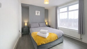 A modern bedroom with a large bed, bedside lamps, a window with curtains, and a city view. - Emerald Living - Swansea (Swansea)