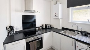 A modern kitchen with a black countertop, stainless steel appliances, white cabinets, a built-in oven, a range hood, a dishwasher, and a window with a view. - Emerald Living - Swansea (Swansea)