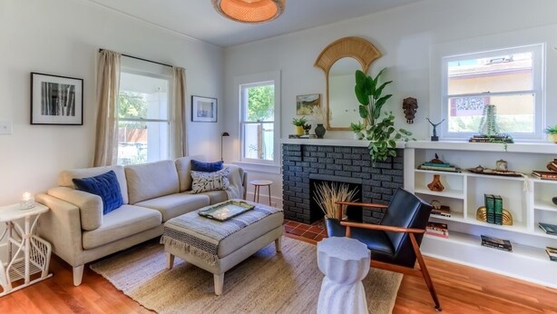 Un salón con sofá, reposapiés y chimenea. Se observan ventanas, un espejo y estantes con objetos decorativos.