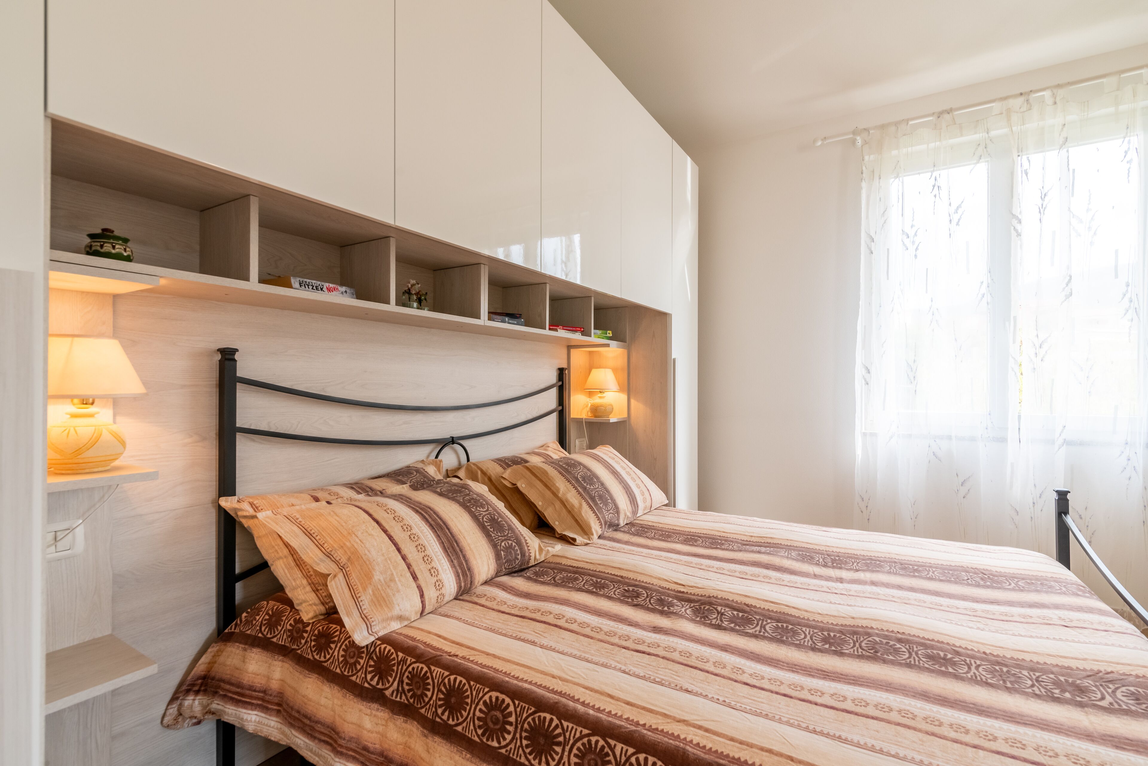 A bedroom with a bed, bedside lamps, a shelf with books, and a window with sheer curtains.