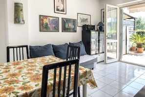 A dining area with a table covered in a sunflower tablecloth, a sofa with blue cushions, and a glass door leading to an outdoor area with potted plants.