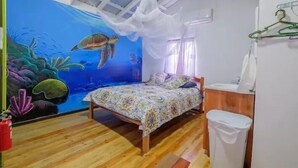 A bedroom with a mural of a sea turtle, a bed with a patterned bedspread, a wooden floor, and a white trash bin. - Mr. Ed's Adventures Belize (San Pedro)