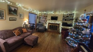 Una sala de estar acogedora con un sofá, un reposapiés y un árbol de Navidad. Hay estanterías, una chimenea y un televisor.